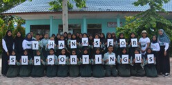 Eine Schulklasse sowie einige Erwachsene stehen vor einem Schulgebäude. Jedes der Kinder hält einen Buchstaben hoch, zusammen ergeben diese Buchstaben „Thanks for supporting us“.