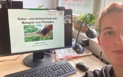Eine Mitarbeiterin lächelt in die Kamera, im Hintergrund zeigt ein Bildschirm eine Zoom-Konferenz.