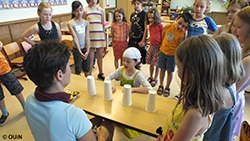 Eine Gruppe von Kindern im Grundschulalter steht um einen Tisch mit Bechern, auf dem ein Spiel gespielt wird.