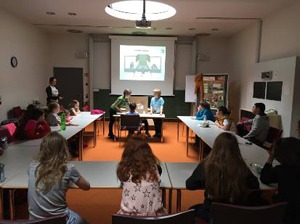 Kinder sitzen an einem Tisch in einem Seminarraum und diskutieren, eine Präsentation wird an eine Leinwand projiziert.