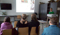 Ein Foto aus der Perspektive eines Zuschauers des Vortrags. Eine Präsentation wird an die Wand projiziert.