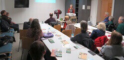 Einige Menschen sitzen an Tischen und hören sich eine Präsentation von Julia Cissewski an.