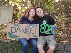 Susann Jänig und Robert Eisenberg sitzen auf einer niedrigen Mauer und halten ein Schild hoch, auf dem „Danke!“ steht, sowie eine aus Pappe ausgeschnittene 800.