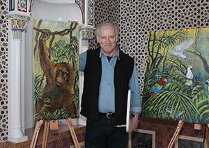 Karl Zwolsky mit zweieni seiner Gemälde. Eines der Bilder zeigt einen Orang-Utan, der sich an einem Ast festhält, und das andere Bild einen Blick in den Regenwald mit einigen Papageien.