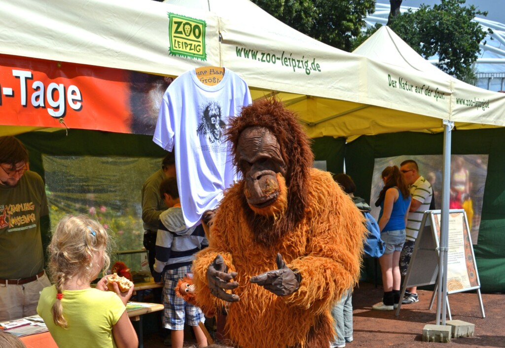 Der Kostüm-Orang-Utan redet vor einem Informationsstand mit einem Kind.
