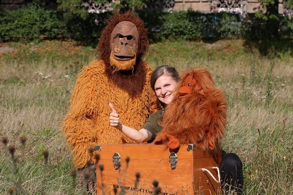 Umweltbildung in Deutschland. Eine Mitarbeiterin des Vereins mit Orang-Utan-Handpuppe „Ginting“ und ein anderer Mitarbeiter im Orang-Utan-Kostüm sitzen auf einer Wiese.