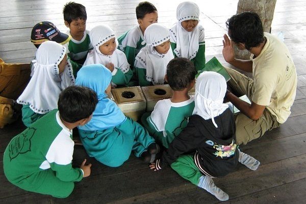 Eine Gruppe kleiner indonesischer Kinder sitzt mit dem Umweltbildner auf dem Holzfußboden und lauscht ihm gespannt.