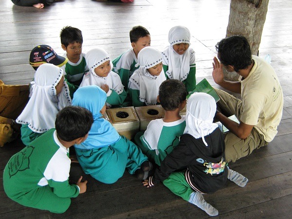 Eine Gruppe kleiner indonesischer Kinder sitzt mit dem Umweltbildner auf dem Holzfußboden und lauscht ihm gespannt.