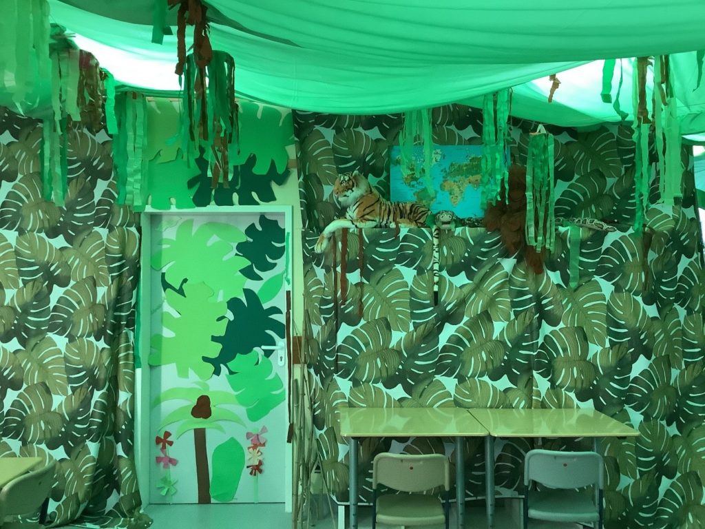 Blick in den Regenwaldraum. Die Wände sind mit Tüchern verkleidet, auf denen Muster aus Blättern gedruckt sind. Die Decke ist mit grünen Tüchern verkleidet. An die Tür wurden aus Pappe gebastelte Blumen und Pflanzenblätter gehängt.