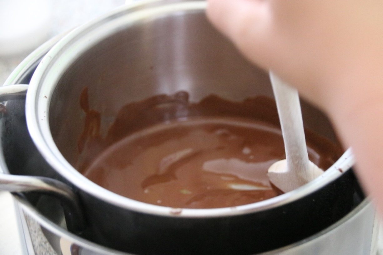 Ein Schüler rührt mit einem Holzlöffel geschmolzene Kuvertüre in einem Topf. Beim Ausprobieren dieses Rezeptes für palmölfreie Schokocreme erfahren die Schüler, dass man leckeren Schokoaufstrich günstig und einfach selbst herstellen kann.