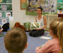 ALT-Text: Die Umweltbildnerin zeigt den im Halbkreis um sie auf dem Boden sitzenden Vorschülern Bildkarten, die die Unterschiede zwischen den Jahreszeiten in Deutschland und im tropischen Regenwald darstellen. Im Hintergrund sieht man weiteres Anschauungsmaterial wie Maskottchen Ginting, Bücher über den Regenwald, getrocknete Kakao-und Kokosfrüchte sowie unser Regenwaldplakat.