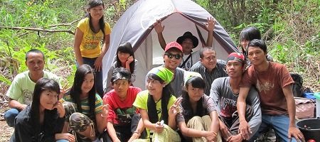 A group of students sit in front of a tent in the forest and look into the camera.