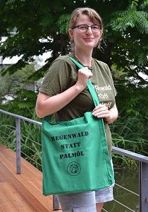 Eine Frau hält den grünen Stoffschulterbeutel mit einem Schulterhenkel. Der schwarze Aufdruck besteht aus „Regenwald statt Palmöl“ und dem Vereinslogo.