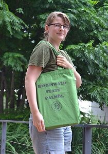 Eine Frau hält den olivgrünen Stoffbeutel mit zwei Schulterhenkeln. Der schwarze Aufdruck besteht aus „Regenwald statt Palmöl“ und dem Vereinslogo.