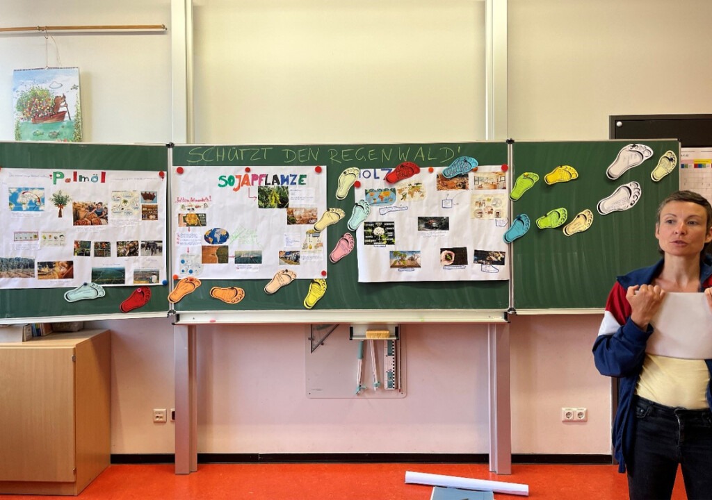 Die Umweltbildnerin steht vor der Klasse. An der Tafel im Hintergrund hängen unter der Überschrift „Schützt den Regenwald“ die drei von den Schülern gestalteten Plakate zu den Themen Palmöl, Soja und Holz. Dazwischen schlängeln sich bunte Fußabdrücke aus Papier.