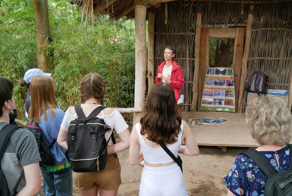 Mehrere Schüler lauschen der Umweltbildnerin vor einer Holzhütte im Zoo Leipzig.