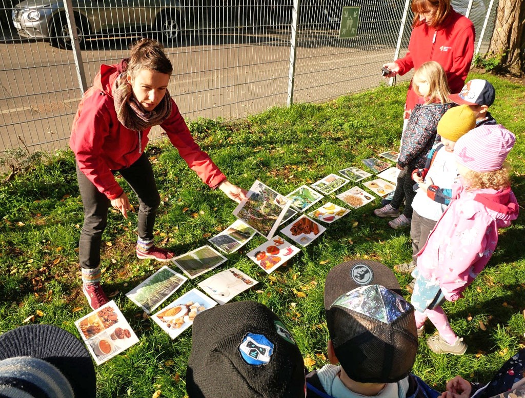Verschiedene Bilder von Alltagsprodukten und Pflanzen aus dem Regenwald liegen auf dem Boden. Eine Gruppe Kinder steht vor den Bildern. Die Umweltbildnerin hält ein Bild hoch und zeigt es der Gruppe.