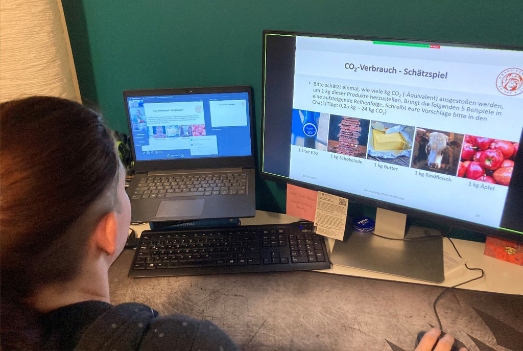 Die Umweltbildnerin schaut auf zwei Monitore. Auf dem linken Monitor sind die Teilnehmenden des Online-Seminars, auf dem rechten Monitor die PowerPoint-Folie zu einem Spiel. Auf der Folie sind Bilder verschiedener Produkte dargestellt, zum Beispiel Äpfel, Rindfleisch oder Butter.