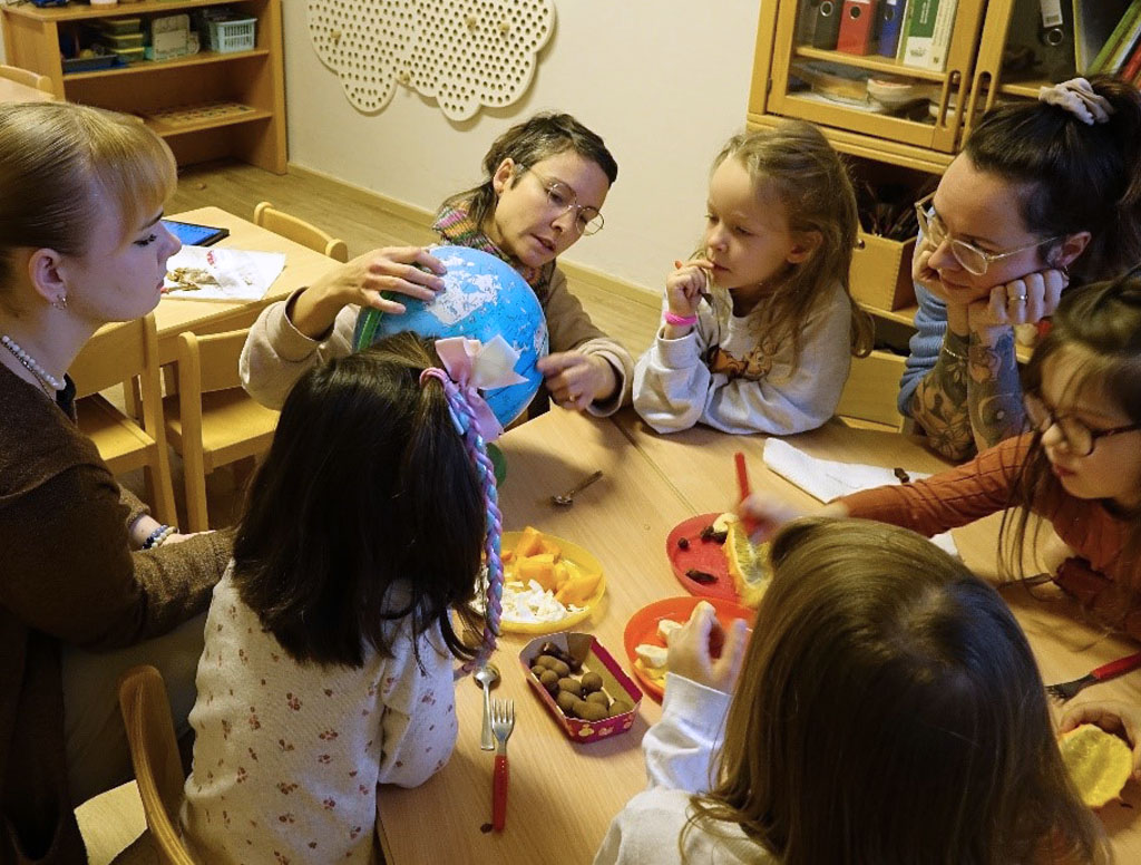 Während sie exotische Früchte kosten, beobachten die Kinder die Umweltbildnerin, die ihnen auf einem Globus die Regenwälder zeigt.