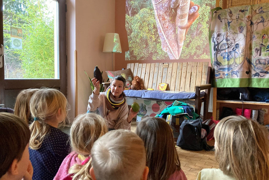 Die Umweltbildnerin zeigt den vor ihr sitzenden Kindern Früchte aus dem Regenwald: eine Kakaoschote und eine Mango. Im Hintergrund hängt ein Plakat mit Regenwaldtieren, ein großes Bild eines kletternden Orang-Utans wird an die Wand projiziert.