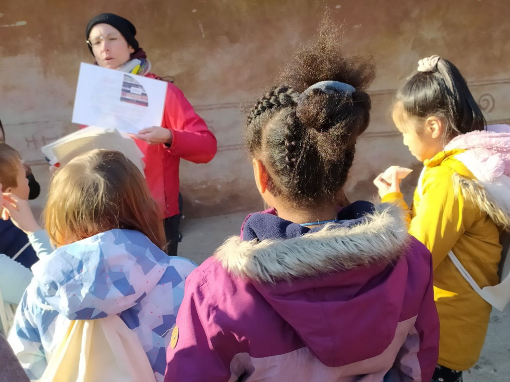 Kinder beobachten die Umweltbildnerin, die ihnen einen Frage stellt und dazu eine Bildkarte mit einer möglichen Antwort zeigt.