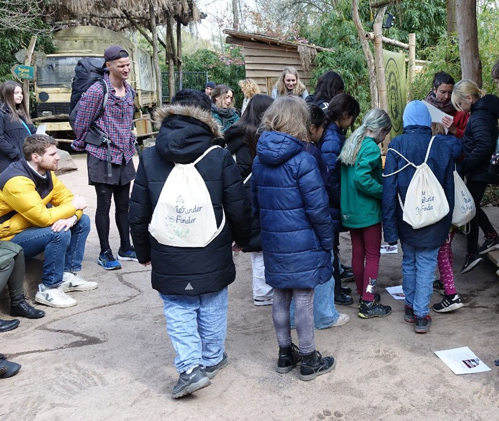 Vor einer Holzhütte ist eine Gruppe Grundschulkinder versammelt. Vor ihnen liegen drei verschiedene Bilder auf dem Boden. Die Kinder spielen das Spiel „Eins, Zwei oder Drei“ und stellen sich zu einem der drei Bilder, auf denen mögliche Antworten zu einer Frage zu sehen sind.