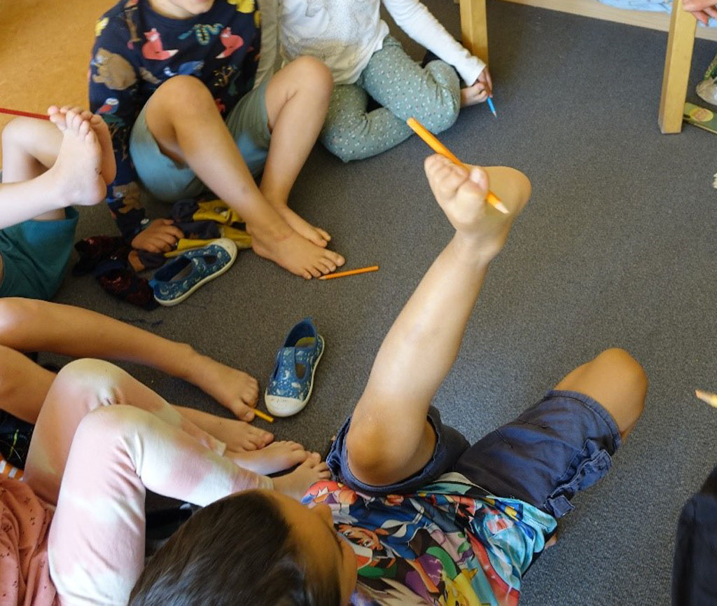 Kinder sitzen im Kreis. Sie sind barfuß und halten Stifte zwischen ihren Zehen.