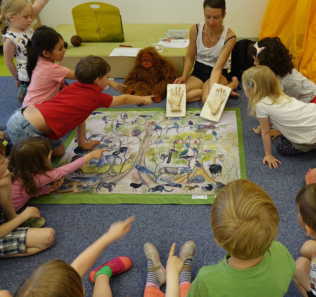 Kinder sitzen im Kreis, in dessen Mitte ein Plakat vom Regenwald und seinen Bewohnern liegt. Die Referentin zeigt den Kindern einen Hand- und einen Fußabdruck eines Orang-Utans. Neben ihr sitzt die Orang-Utan-Handpuppe Ginting.