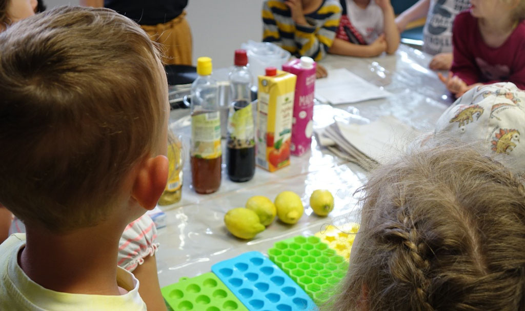 Kinder stehen um einen Tisch. Auf diesem befinden sich verschiedene Säfte, Zitronen, Agavensaft und Silikonförmchen für die Herstellung palmölfreier Gummibärchen.