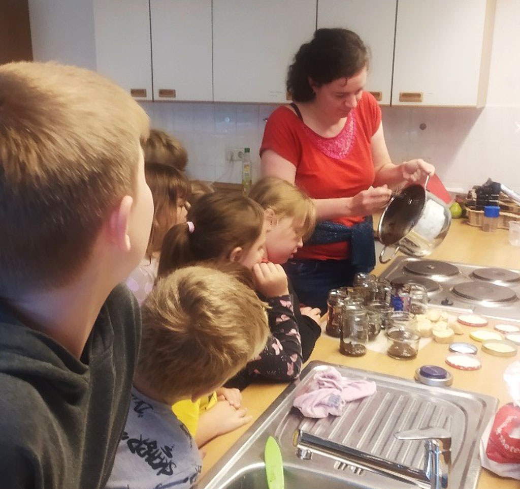 Kinder stehen am Herd und schauen zu, wie eine Frau Schokocreme aus einem Topf in Gläser abfüllt.