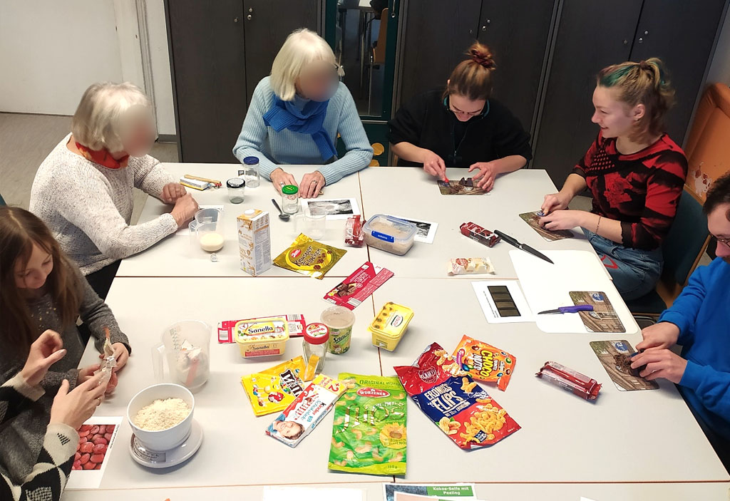 An einem Tisch sitzen Workshop-Teilnehmende. Sie wiegen gemahlene Nüsse, schneiden Schokolade und messen Milch für die Zubereitung einer Schokocreme ab. Auf dem Tisch liegen verschiedene Verpackungen von Lebensmitteln mit und ohne Palmöl.