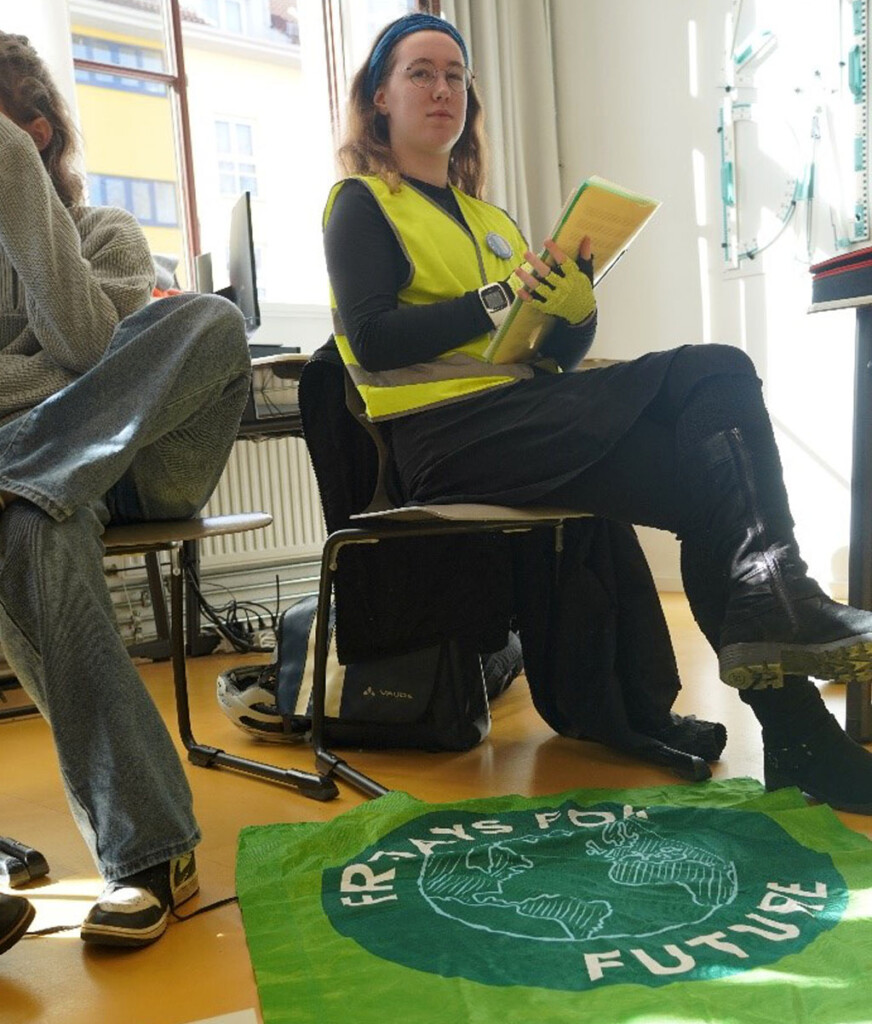 Im Vordergrund liegt ein grünes Fridays for Future Plakat auf dem Boden, dahinter sitzt eine Schülerin, sie trägt eine Warnweste und hält Unerlagen im Arm. Am linken Bildrand ist eine weitere Person angeschnitten.