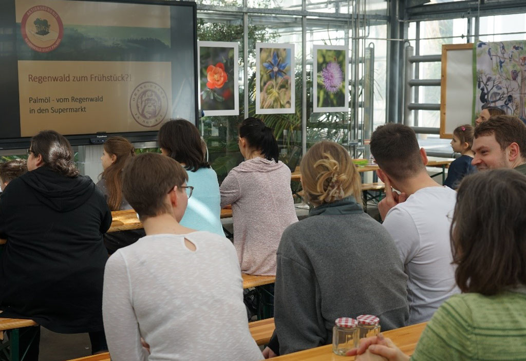 Erwachsene und Kinder sitzen auf Bänken und schauen auf eine Beamer-Präsentation. Daneben sind Bilder von Pflanzen zu sehen.