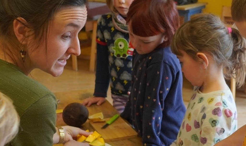 Auf der rechten Bildhälfte stehen vier Kinder an einem Tisch, auf dem Schneidebretter und -messer, Bananenschalen, eine Kakaobohne und eine Kokosnuss liegen. Die Umweltbildnerin auf der linken Seite schneidet Mango auf.