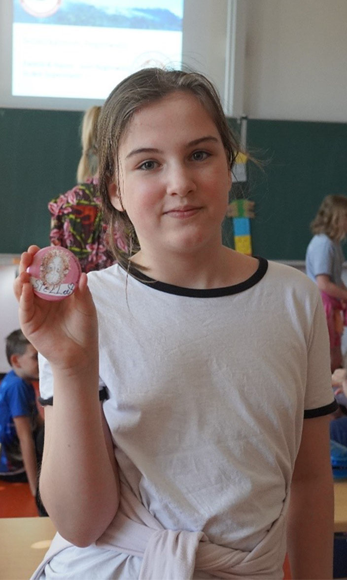 Ein Kind hält einen Button in der Hand, darauf steht sein Name. Im Hintergrund sind weitere Kinder und eine Schultafel sowie eine Präsentation.