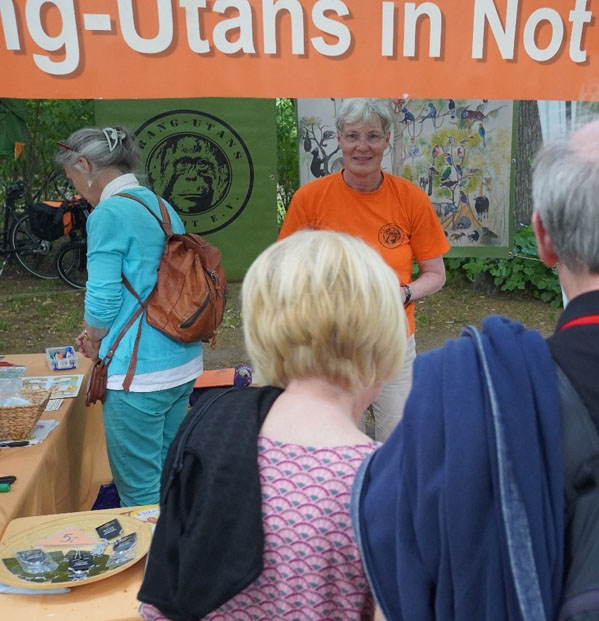 Zwei Personen stehen vor dem Stand von Orang-Utans in Not e.V. Am rechten Bildrand befinden sich ein grünes T-Shirt und ein Plüsch-Orang-Utan. Hinter dem Stand stehen ebenfalls zwei Personen.