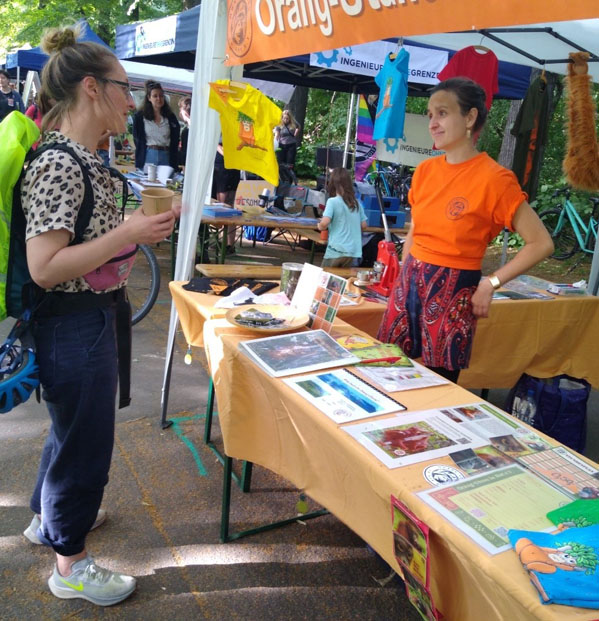 Vor und hinter einem Informationsstand steht jeweils eine Person. Es liegen verschiedene Flyer und Materialien aus, an einem Pavillion sind T-Shirts und ein Plüsch-Orang-Utan befestigt. Im Hintergrund befinden sich weitere Informationsstände und Personen.