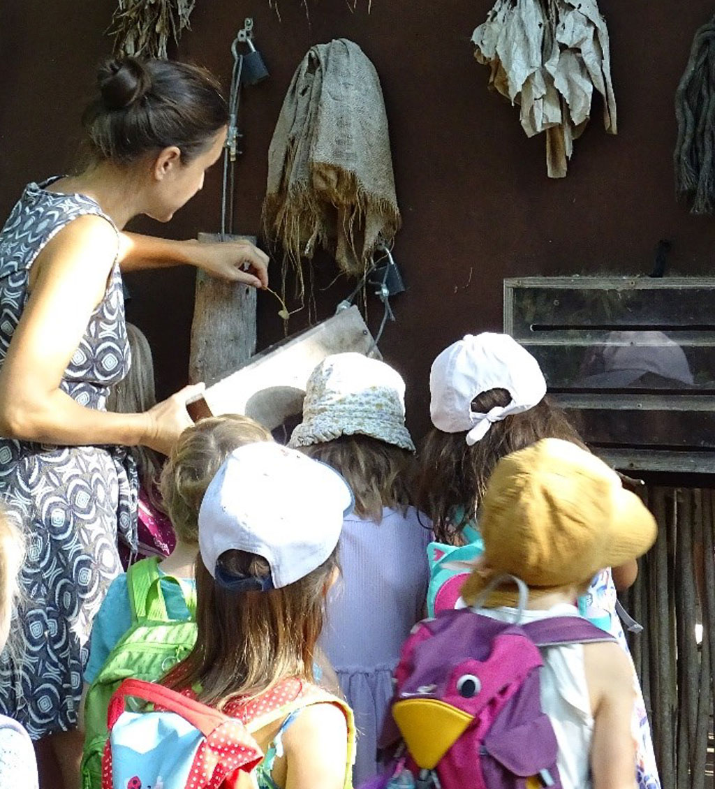 Die Umweltbildnerin steht vor einer Schautafel mit verschiedenen Beschäftigungsmaterialien für Menschenaffen. Eine Gruppe Kinder befindet sich im Vordergrund.