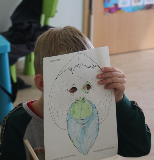 Ein Kind hält sich ein Blatt Papier vor sein Gesicht. Darauf ist die Maske eines männlichen Orang-Utans abgebildet.