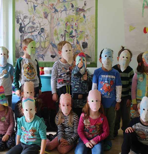 Mehrere Kinder hocken und stehen vor einem Regenwaldplakat. Alle tragen Orang-Utan-Masken.