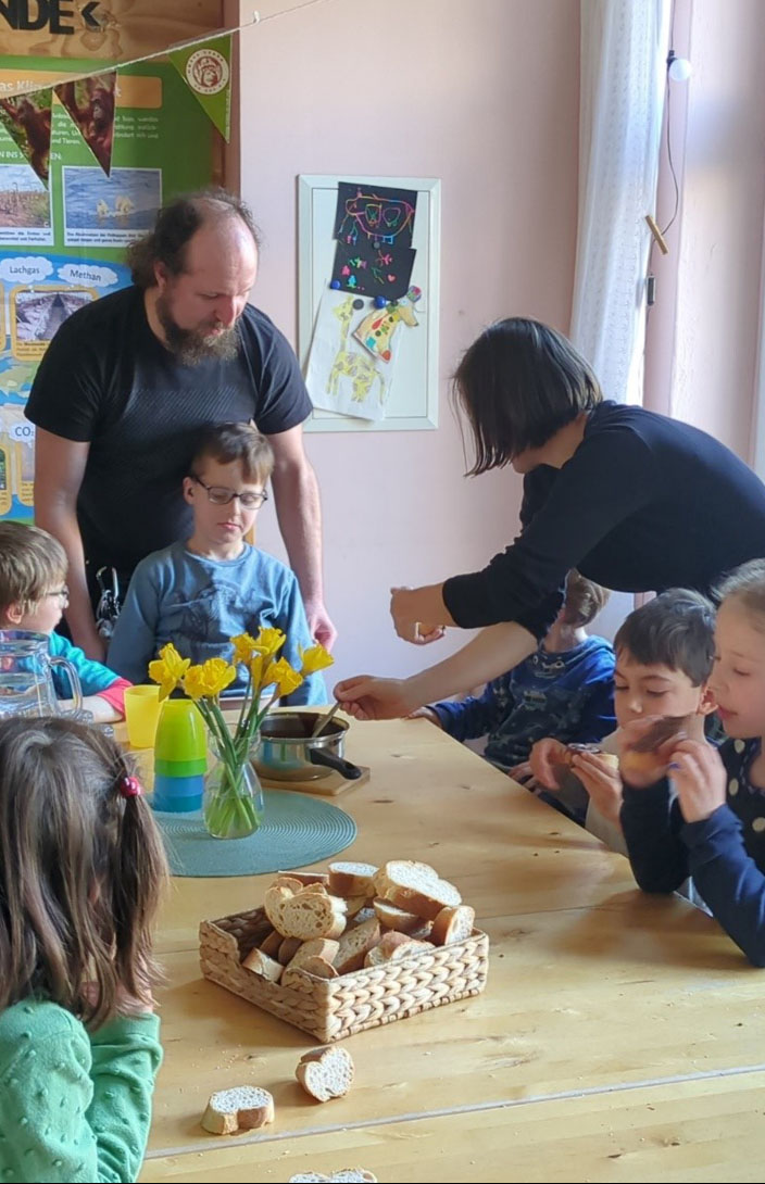 Kinder sitzen an einem Tisch, auf dem ein Brotkorb mit aufgeschnittenem Brot steht. Weiter befindet sich darauf eine Vase mit gelben Blumen, verschiedenfarbige Becher und ein Topf aus dem die Umweltbildnerin mit einem Löffel schöpft.