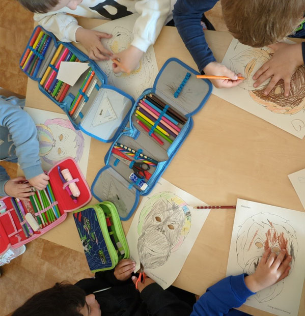 Kinder sitzen um einen Tisch herum. Darauf liegen Ausmalbilder von Orang-Utan-Masken und Buntstifte.