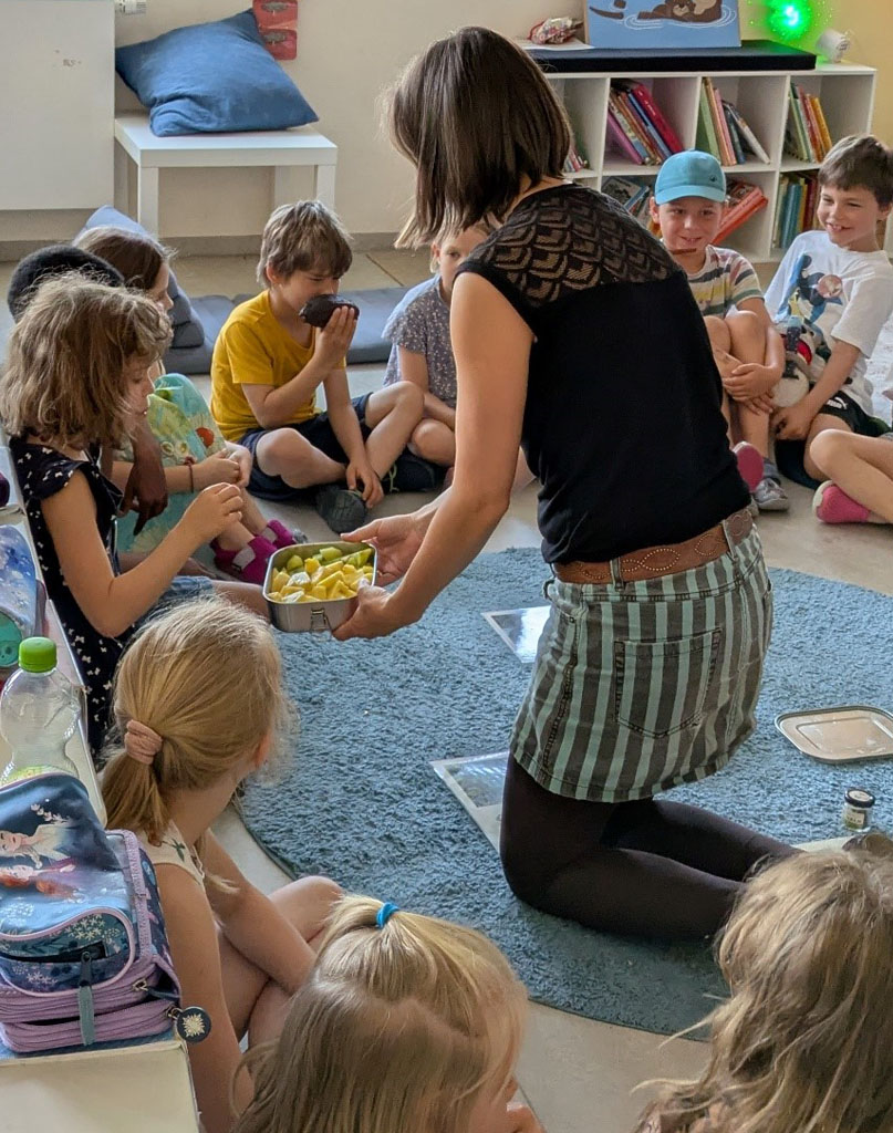 Kinder sitzen im Kreis auf dem Boden. Die Umweltbildnerin teilt Obst aus. Ein Kind riecht an einer Kakaofrucht.