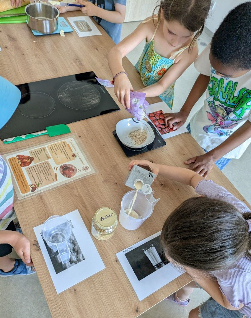 Kinder stehen um eine Kücheninsel herum. Ein Kind füllt Milch in einen Messbecher. Zwei Kinder wiegen gemahlene Nüsse ab.