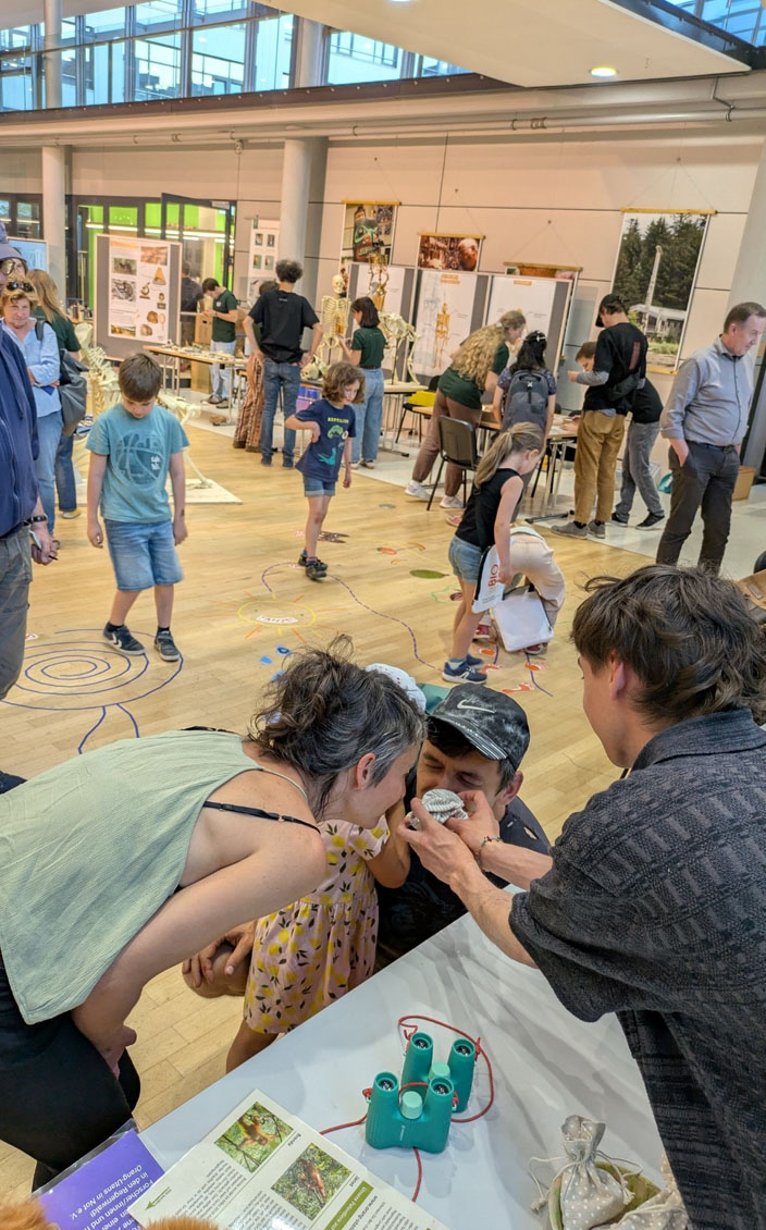 In der Eingangshalle des Max-Planck-Instituts für evolutionäre Anthropologie sind viele Besucher und verschiedene Stationen. Auf dem Boden ist ein farbiger Parcours aus Bildern und Klebestreifen aufgebracht, der von verschiedenen Personen absolviert wird. Im Vordergrund des Bilds steht eine Familie und riecht in einen Sack hinein. Auf dem Tisch liegen Ferngläser, weitere Säckchen und ein Informationsblatt.