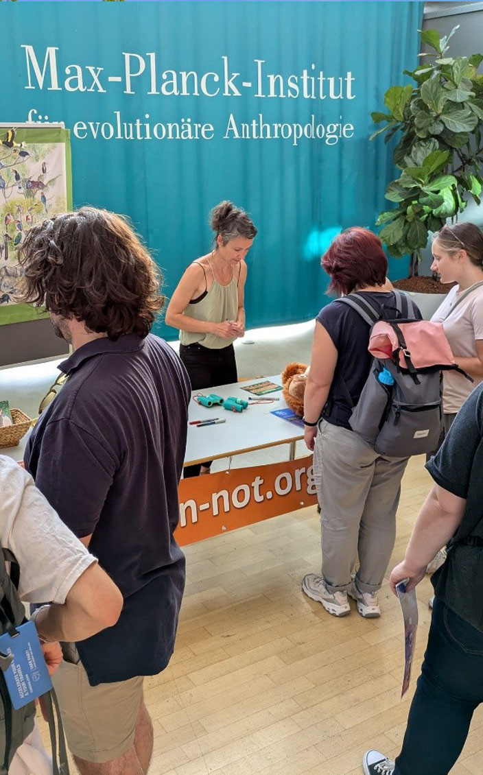 Besucher stehen vor dem Stand des Orang-Utans in Not e.V. Dahinter steht eine Frau. Im Hintergrund hängt ein Plakat sowie eine Regenwald-Girlande vor dem Vorhang des Max-Plack-Instituts.