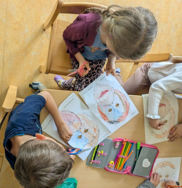 Kinder sitzen an einem Tisch und malen Orang-Utan Masken bunt aus. Ein Kind schneidet gerade seine Maske aus.