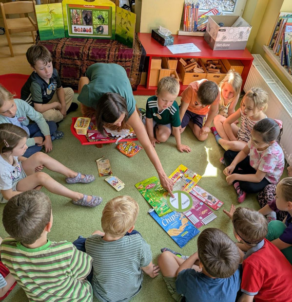 Kinder sitzen im Kreis auf dem Boden. In der Mitte liegen verschiedene Produktverpackungen. Die Umweltbildnerin zeigt auf eine Verpackung.