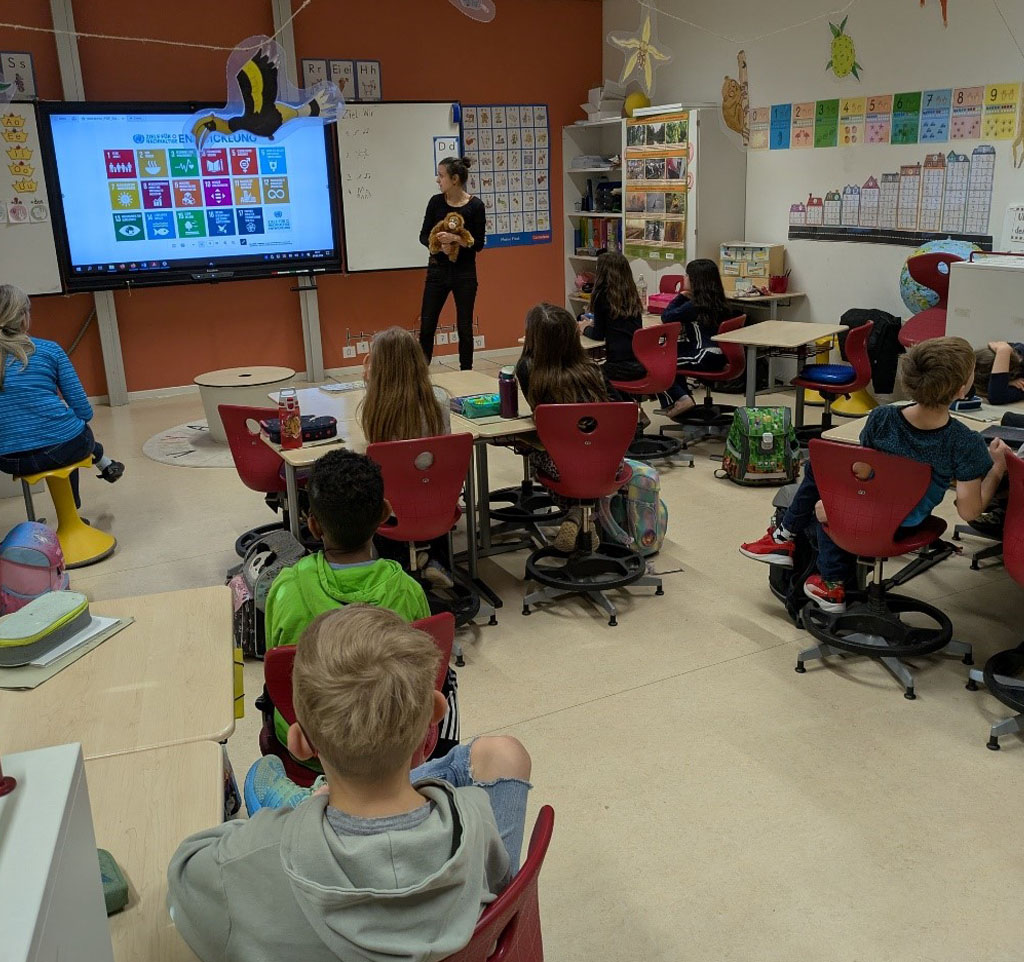Im Klassenzimmer sitzen Kinder an Tischen und schauen der Umweltbildnerin zu, die an einer digitalen Tafel steht und einen Plüsch Orang-Utan in der Hand hält. Am oberen Bildrand hängt eine Regewald-Girlande, bestehend aus einem Hornvogel, einer Schlange, einem Koboldmaki, einer Vanilleorchideenblüte, einer Durianfrucht und einem Orang-Utan.