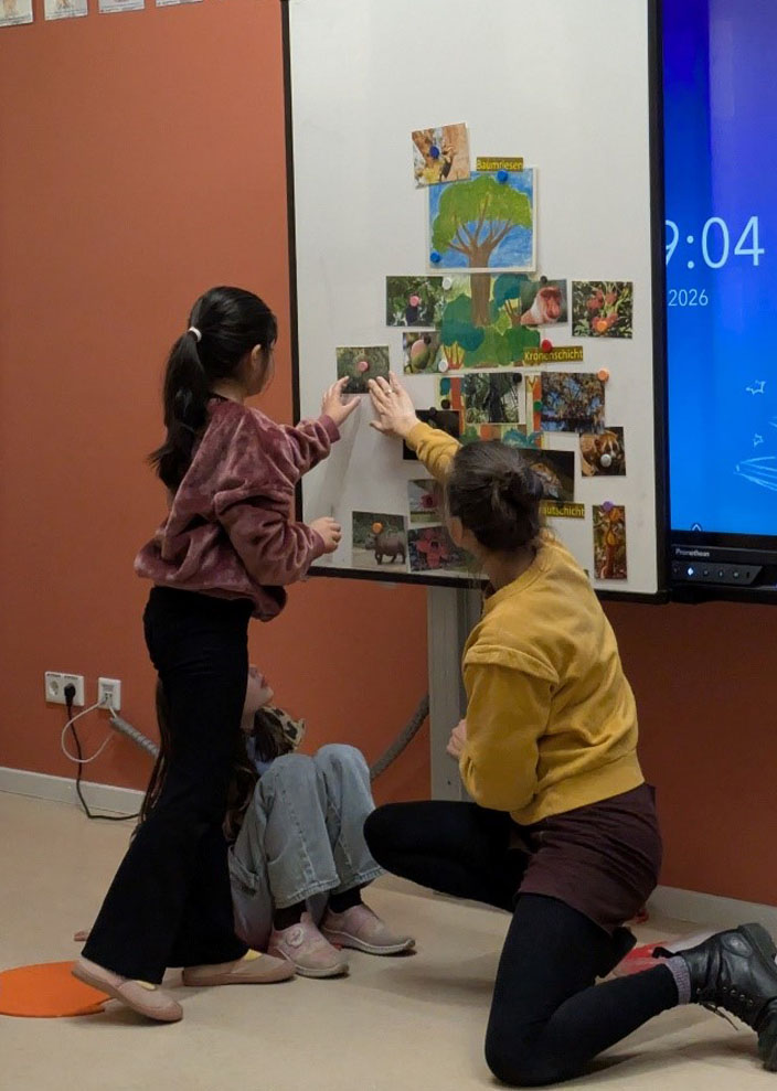 Ein Kind befestigt ein Bild an der Tafel, an der schon andere Bilder angebracht sind. Die Umweltbildnerin unterstützt das Kind dabei.